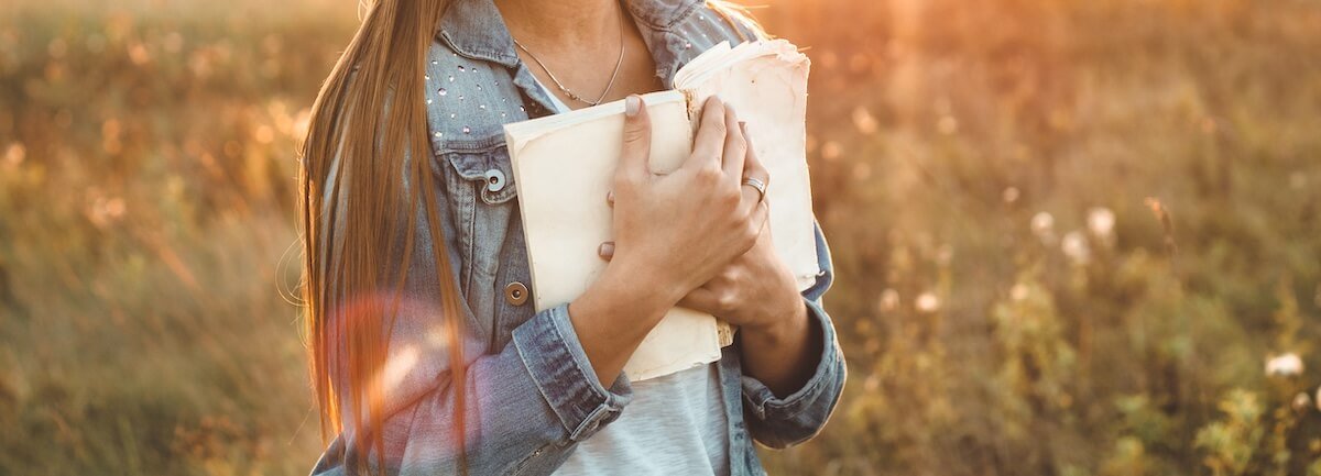 Woman standing in a sunlit field holding a Bible close to her chest, symbolizing peace, trust, and the comforting care of Jesus.