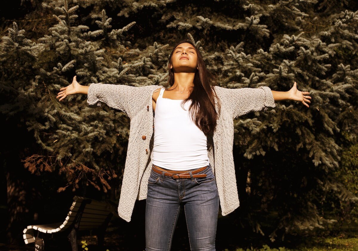 Woman standing outdoors with arms outstretched and eyes closed, embracing sunlight and freedom — symbolizing spiritual healing, surrender, and freedom from past struggles.