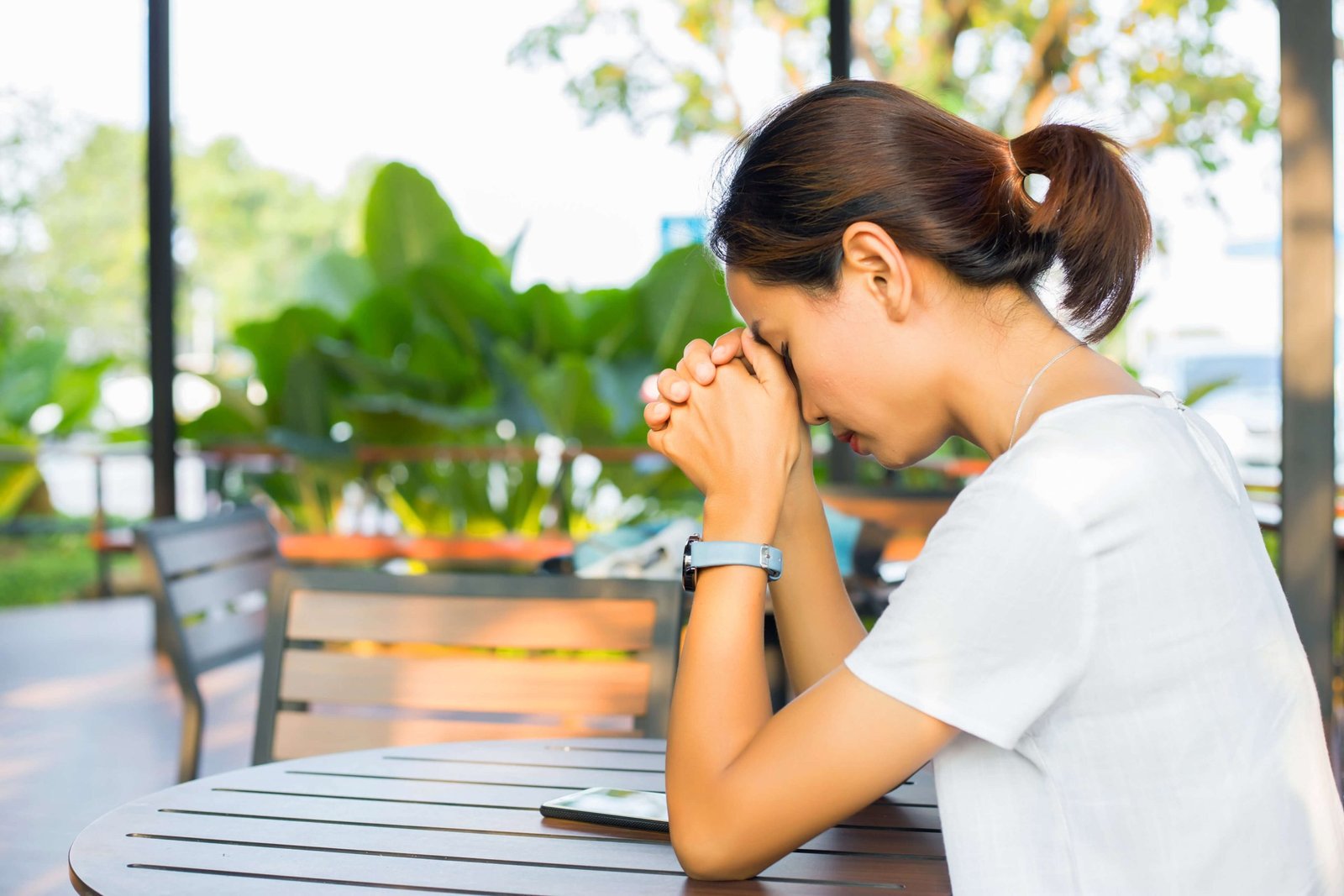Woman sitting at an outdoor table with her head bowed and hands clasped in prayer, symbolizing peace, faith, and finding calm in the midst of a busy day.