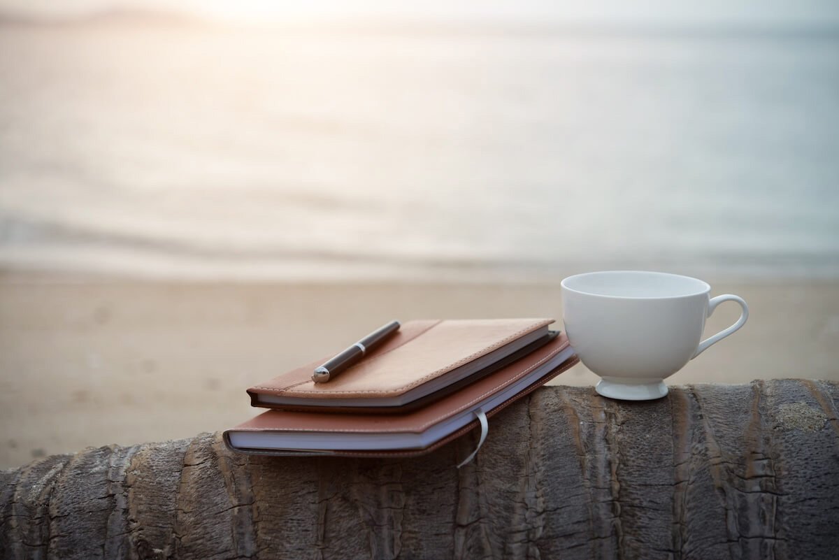 Journal and coffee cup resting on a wooden surface near the beach, symbolizing quiet reflection, Sabbath rest, and slowing down to hear God’s voice.