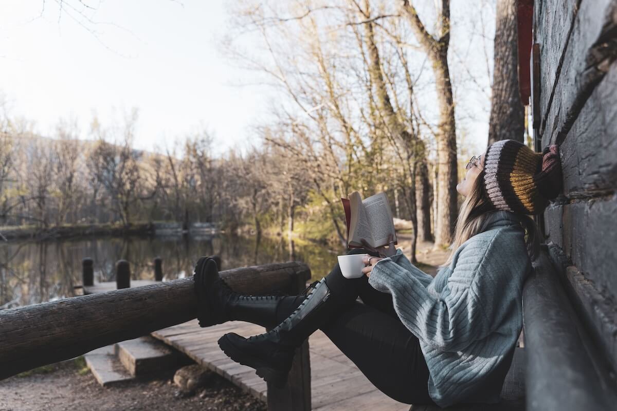 Young woman sitting outside a wooden cabin with a cup of coffee and an open book, symbolizing Sabbath rest, reflection, and finding peace in God’s presence.