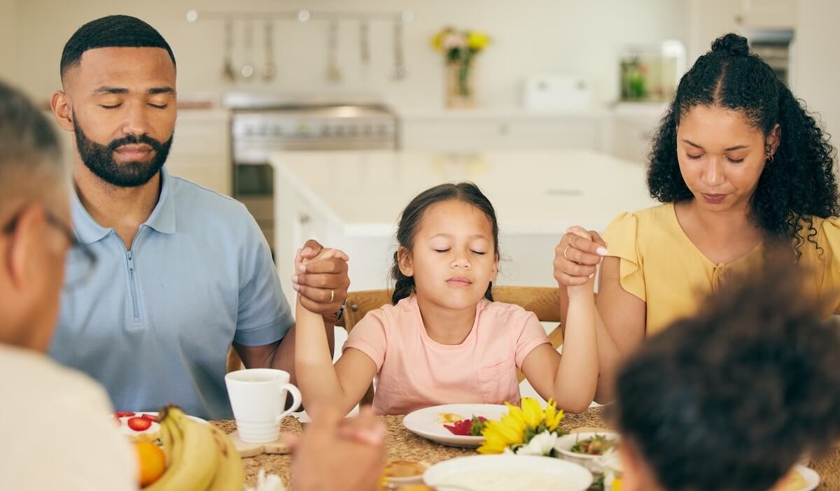 “Family holding hands and praying together at the dinner table, eyes closed, giving thanks before a meal.”